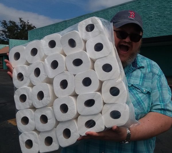 An stunningly beautiful bearded man holds a big bag of toilet tissue and smiles